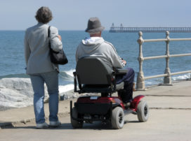 Elderly man riding motorized wheelchair with woman on seaside promenade.