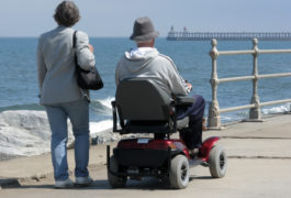 Elderly man riding motorized wheelchair with woman on seaside promenade.